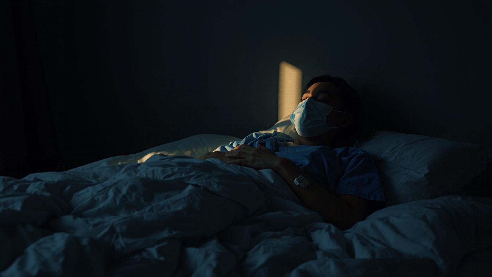 A worker still in blue scrubs sleeps in a darkened bedroom during the day while a narrow strip of sunlight falls on the wall behind the bed
