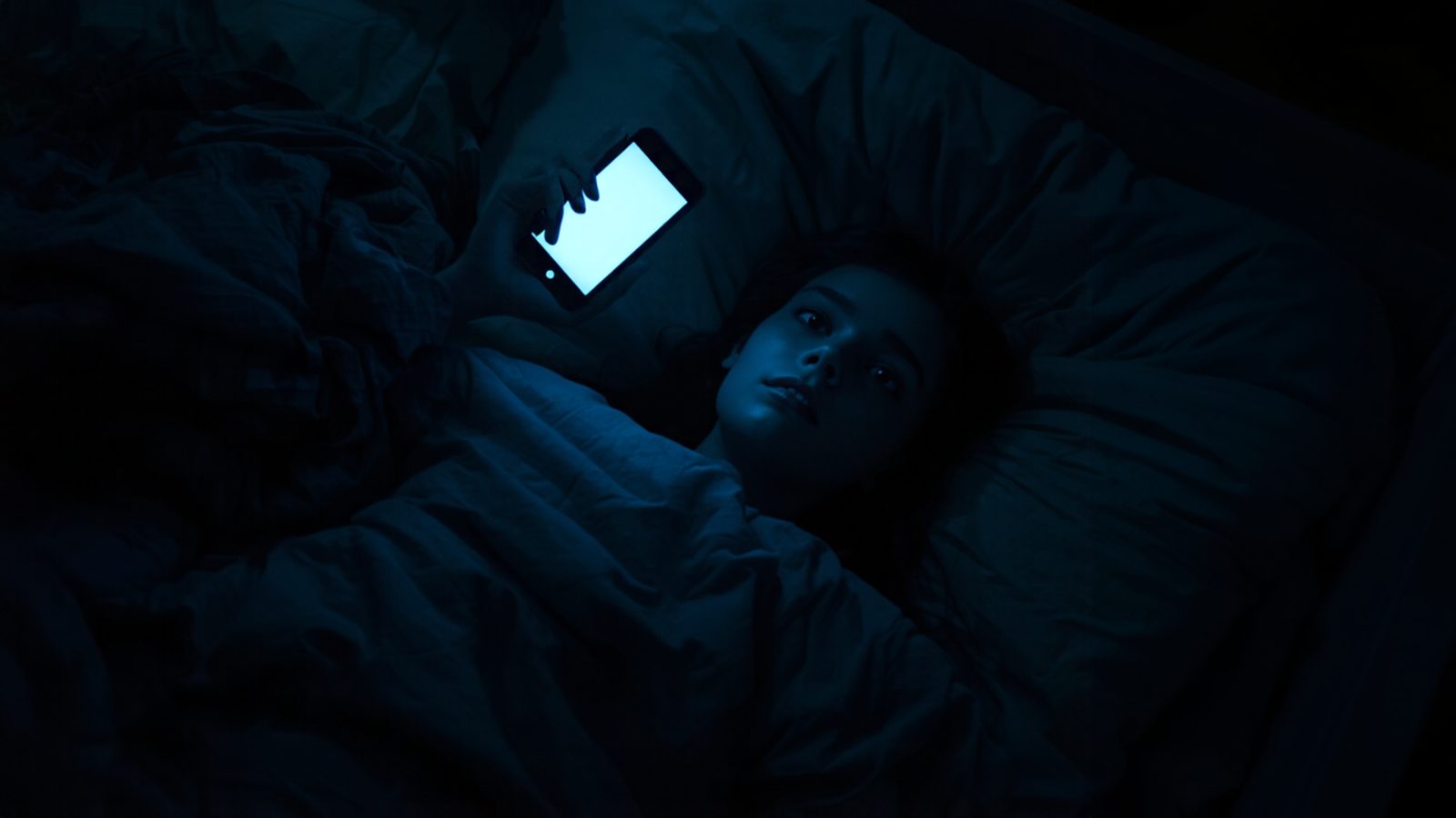 A teenage girl lies awake in bed in a dark bedroom, her face lit by the glow of the smartphone she holds above her pillow